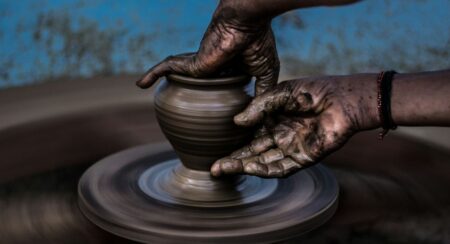 Two hands of a person with dark skin tone shaping bronze-colored clay on a pottery wheel in front of a blurred blue background with brown spots