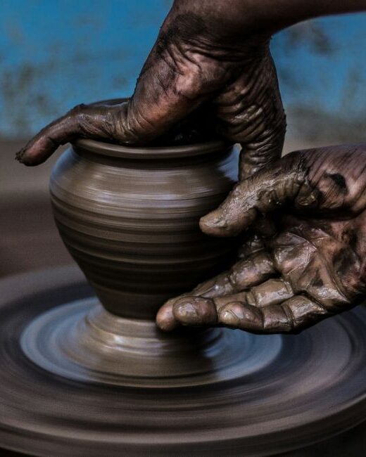 Two hands of a person with dark skin tone shaping bronze-colored clay on a pottery wheel in front of a blurred blue background with brown spots