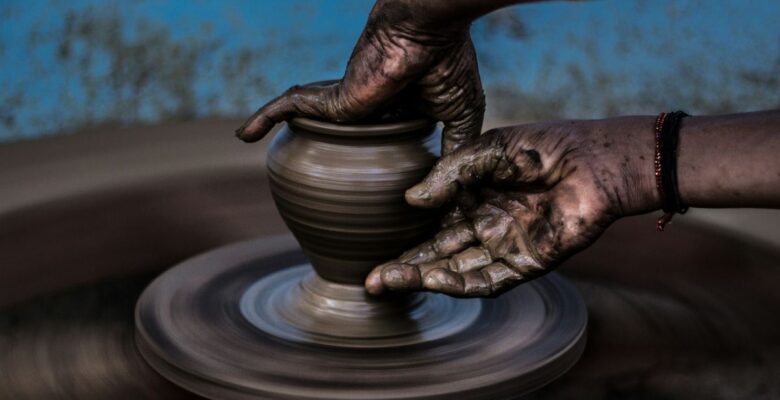 Two hands of a person with dark skin tone shaping bronze-colored clay on a pottery wheel in front of a blurred blue background with brown spots