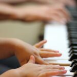 Close-up of the hands and arms of a child with medium skin tone playing piano with the hands of an older person with medium dark skin tone playing in the blurred background