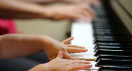 Close-up of the hands and arms of a child with medium skin tone playing piano with the hands of an older person with medium dark skin tone playing in the blurred background