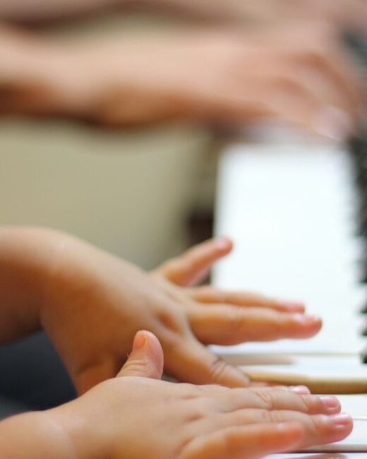 Close-up of the hands and arms of a child with medium skin tone playing piano with the hands of an older person with medium dark skin tone playing in the blurred background