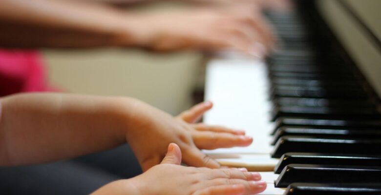Close-up of the hands and arms of a child with medium skin tone playing piano with the hands of an older person with medium dark skin tone playing in the blurred background