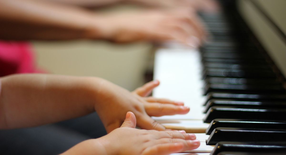 Close-up of the hands and arms of a child with medium skin tone playing piano with the hands of an older person with medium dark skin tone playing in the blurred background