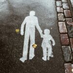 A white painted illustration on black pavement of two human figures, one adult and one child, holding hands; to the right of the figures are two vertical rows of bricks in red, gray, brown, and black