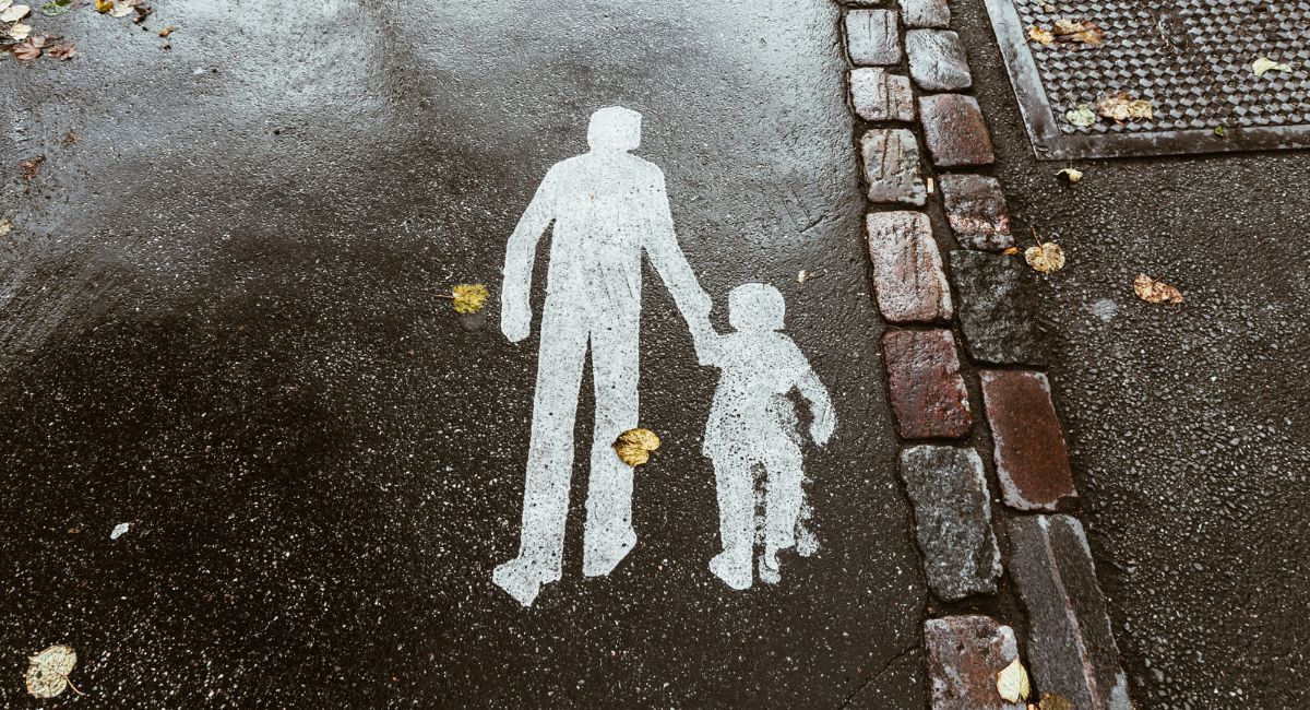 A white painted illustration on black pavement of two human figures, one adult and one child, holding hands; to the right of the figures are two vertical rows of bricks in red, gray, brown, and black