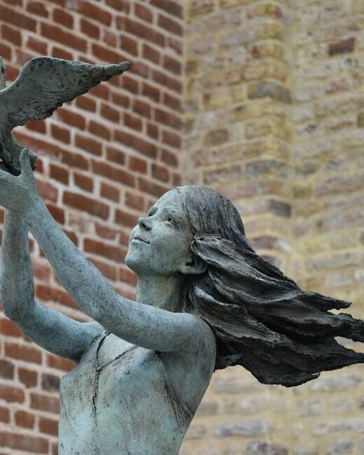 Close-up of a copper statue of a human figure that appears to be a woman with long hair releasing a bird with wings outstretched