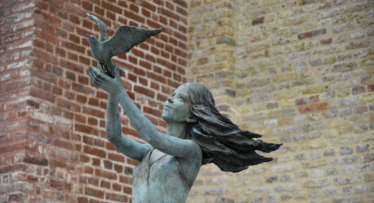 Close-up of a copper statue of a human figure that appears to be a woman with long hair releasing a bird with wings outstretched