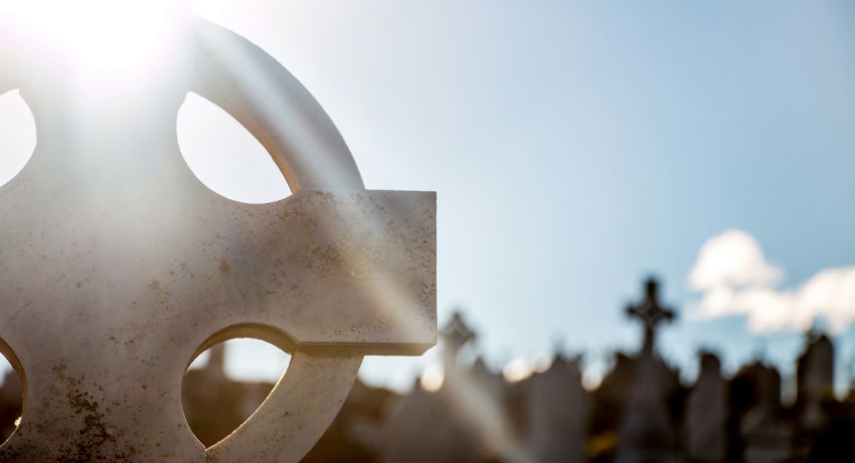 Close-up of sunlit Celtic cross headstone in a cemetery with a blurred background and blue sky