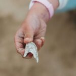 Close-up of the hand of a young person with medium skin tone wearing a pink wristband, holding a gray and white shell in front of a blurred beige background
