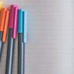 Close-up of four black pens with yellow, orange, pink, and blue caps on an open lined blank notebook