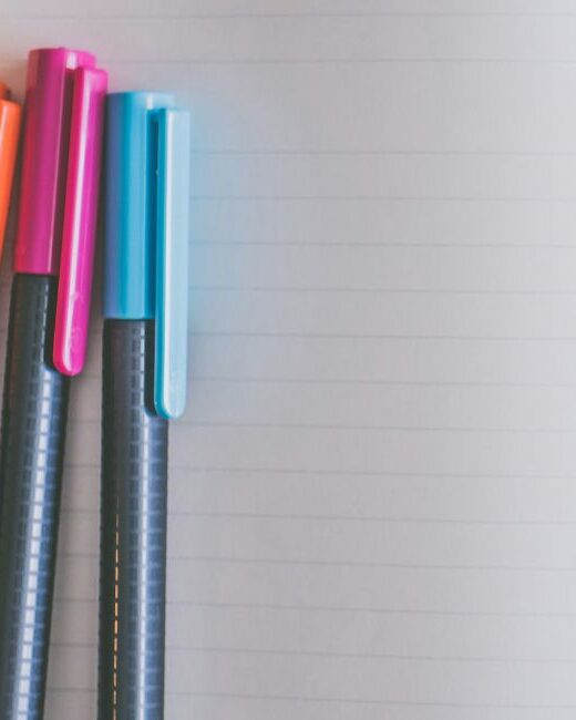 Close-up of four black pens with yellow, orange, pink, and blue caps on an open lined blank notebook