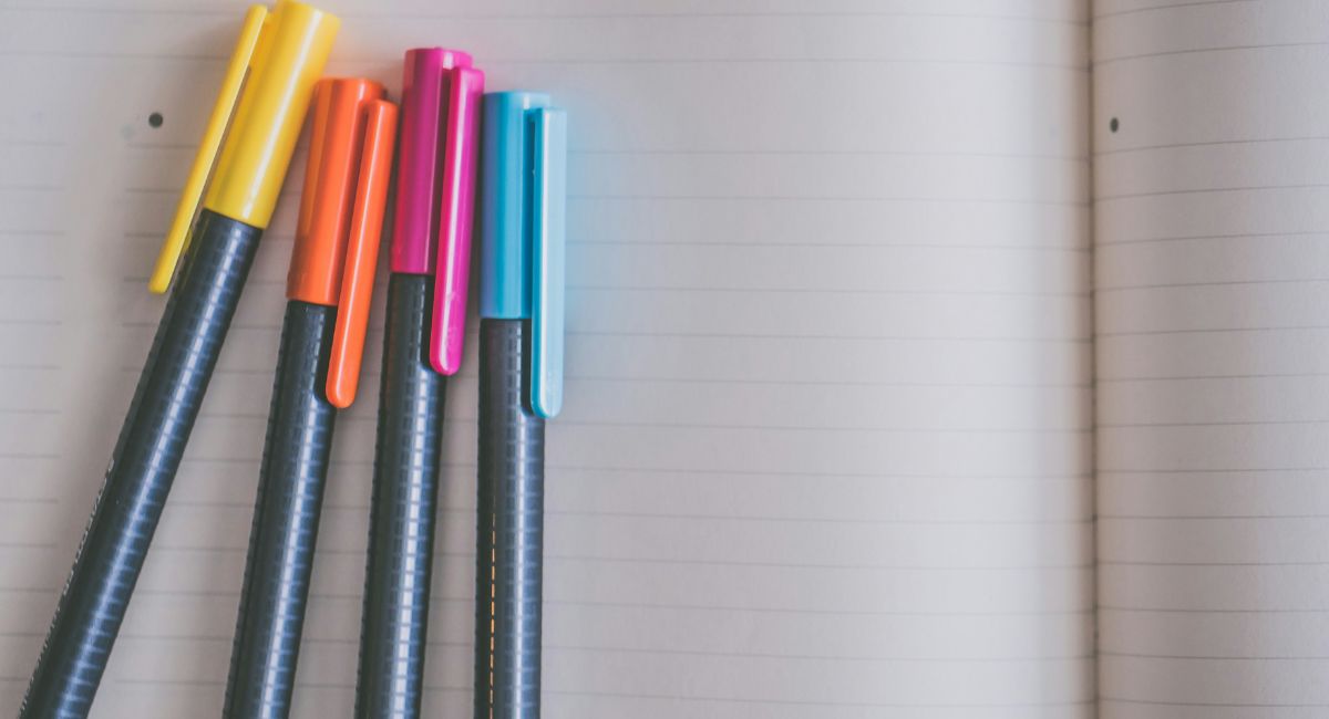 Close-up of four black pens with yellow, orange, pink, and blue caps on an open lined blank notebook