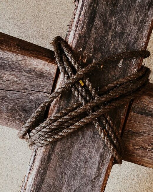 Close-up of dark brown wooden cross with dark brown rope wrapped diagonally over cross beams in front of a light brown background