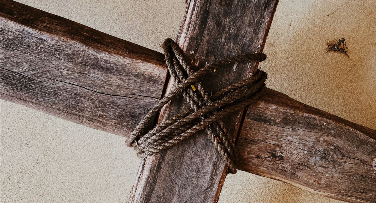 Close-up of dark brown wooden cross with dark brown rope wrapped diagonally over cross beams in front of a light brown background