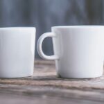 Two white ceramic mugs with handles on a brown wooden surface in front of a blue-gray background