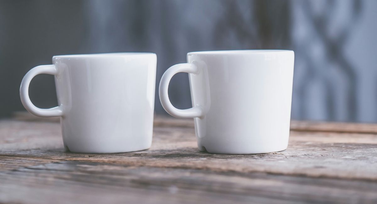 Two white ceramic mugs with handles on a brown wooden surface in front of a blue-gray background