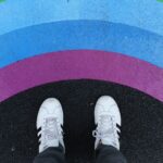 Aerial view of the white and black shoes of a person standing on black pavement before painted arcs in rainbow colors of yellow, green, blue, and purple