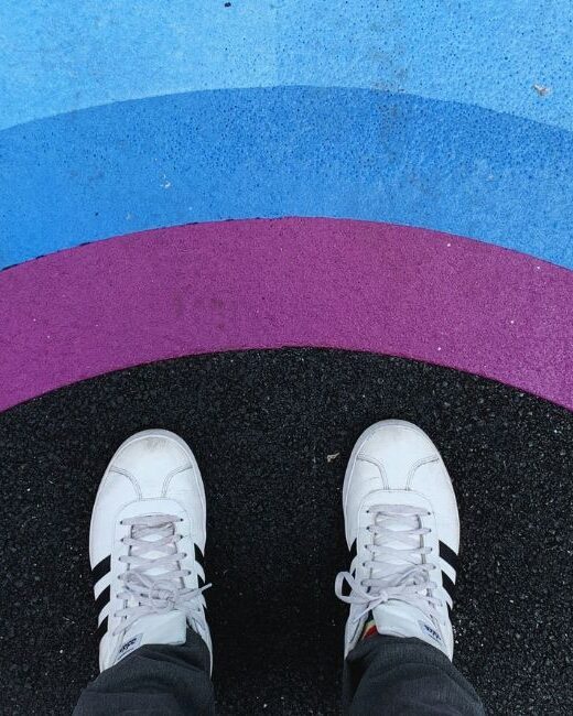 Aerial view of the white and black shoes of a person standing on black pavement before painted arcs in rainbow colors of yellow, green, blue, and purple