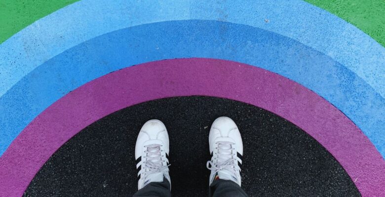 Aerial view of the white and black shoes of a person standing on black pavement before painted arcs in rainbow colors of yellow, green, blue, and purple