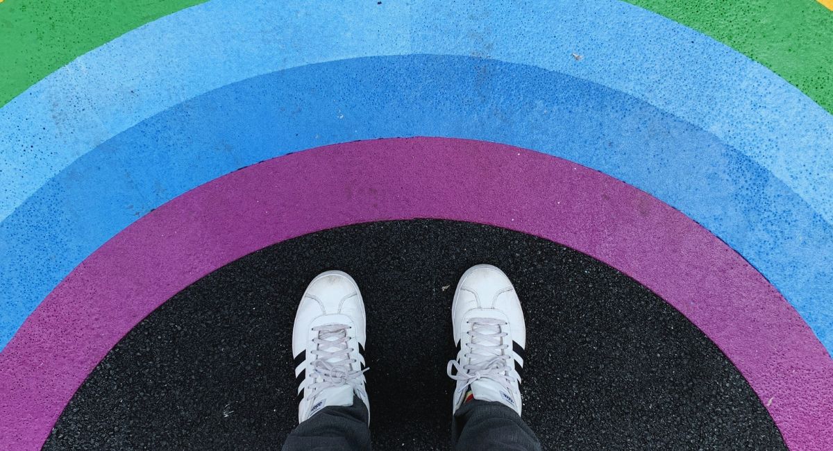 Aerial view of the white and black shoes of a person standing on black pavement before painted arcs in rainbow colors of yellow, green, blue, and purple