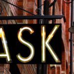Black sign with letters lit up that say "ASK," attached to a red brick building