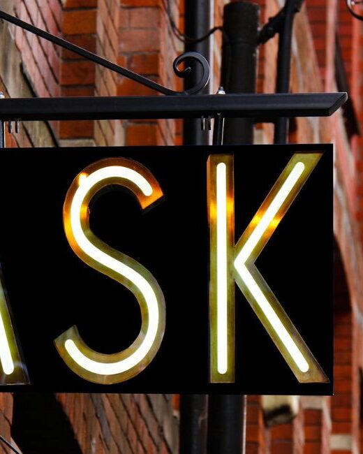 Black sign with letters lit up that say "ASK," attached to a red brick building