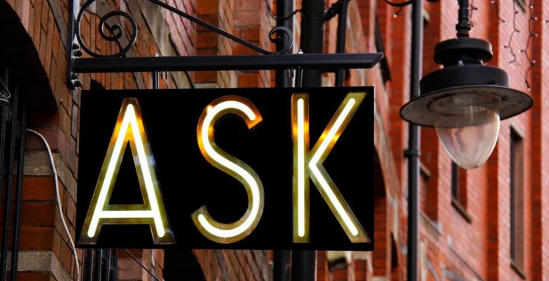 Black sign with letters lit up that say "ASK," attached to a red brick building
