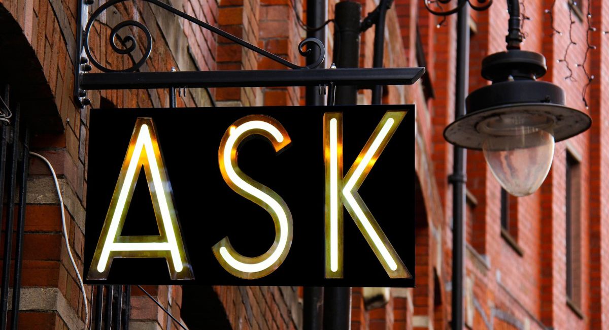 Black sign with letters lit up that say "ASK," attached to a red brick building