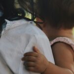 View from the back of an adult with long black hair wearing a white shirt holding an infant with short brown hair and medium dark skin tone wearing a pink and white gingham outfit