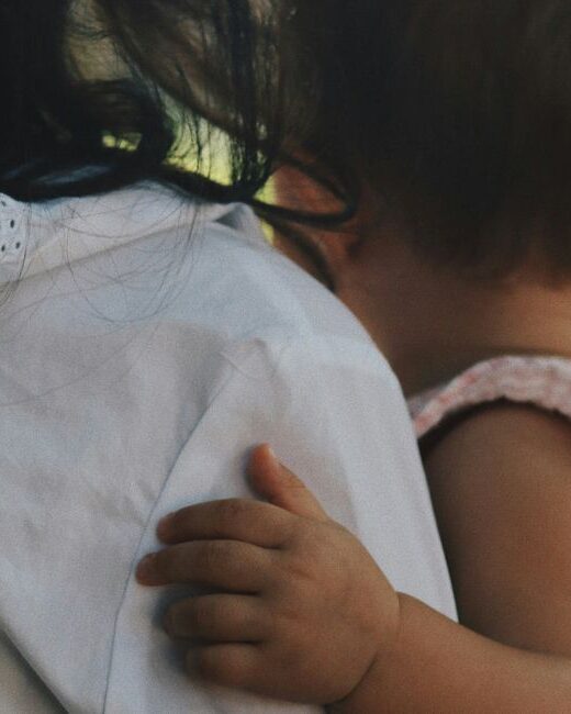 View from the back of an adult with long black hair wearing a white shirt holding an infant with short brown hair and medium dark skin tone wearing a pink and white gingham outfit