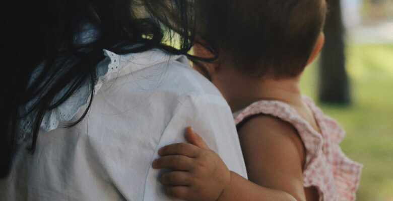 View from the back of an adult with long black hair wearing a white shirt holding an infant with short brown hair and medium dark skin tone wearing a pink and white gingham outfit