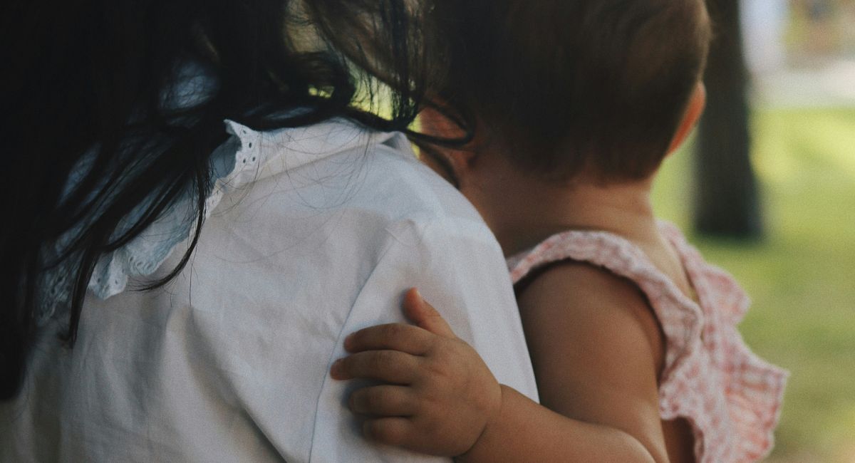 View from the back of an adult with long black hair wearing a white shirt holding an infant with short brown hair and medium dark skin tone wearing a pink and white gingham outfit