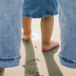 Close-up view from behind of the bare feet of an adult with light skin tone wearing light blue jeans and standing on sand behind the bare feet of a child with medium skin tone wearing a dark blue jean skirt