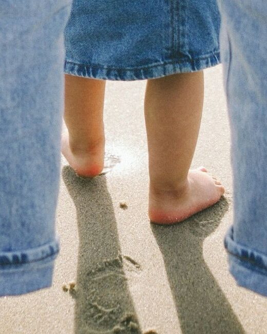 Close-up view from behind of the bare feet of an adult with light skin tone wearing light blue jeans and standing on sand behind the bare feet of a child with medium skin tone wearing a dark blue jean skirt
