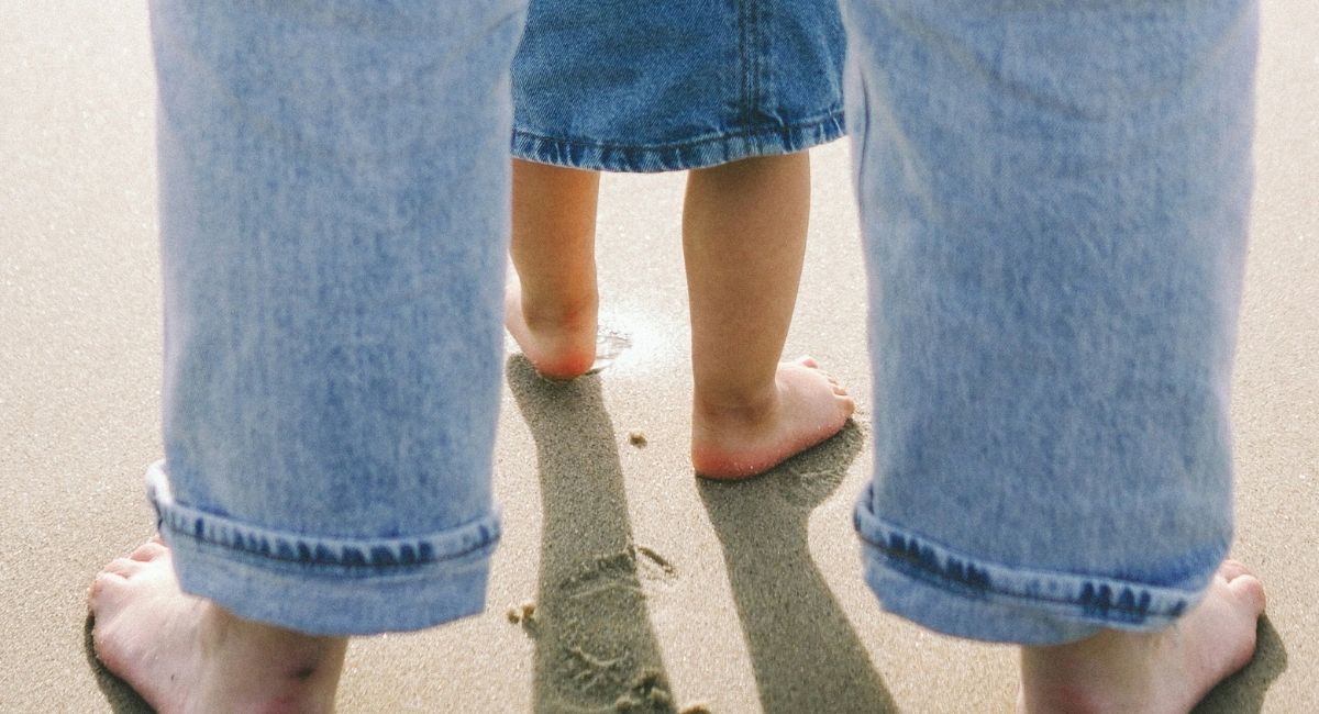 Close-up view from behind of the bare feet of an adult with light skin tone wearing light blue jeans and standing on sand behind the bare feet of a child with medium skin tone wearing a dark blue jean skirt