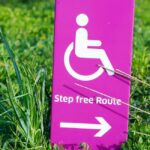 Pink sign in green grass showing (from top to bottom) white wheelchair accessible symbol above text that says "Step free Route" above arrow pointing right