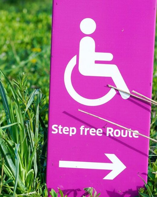 Pink sign in green grass showing (from top to bottom) white wheelchair accessible symbol above text that says "Step free Route" above arrow pointing right