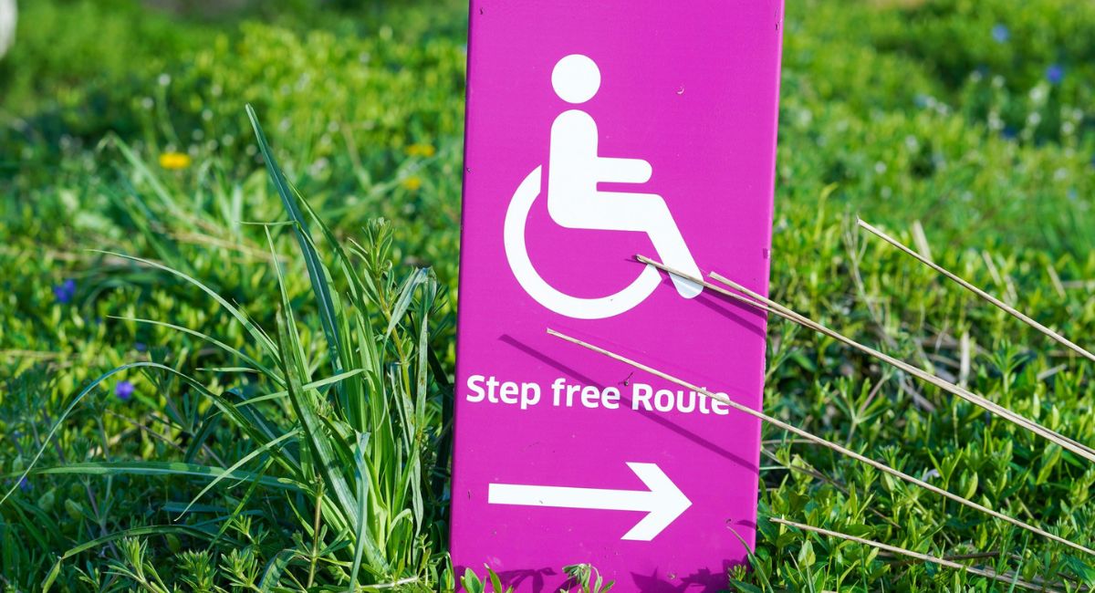 Pink sign in green grass showing (from top to bottom) white wheelchair accessible symbol above text that says "Step free Route" above arrow pointing right